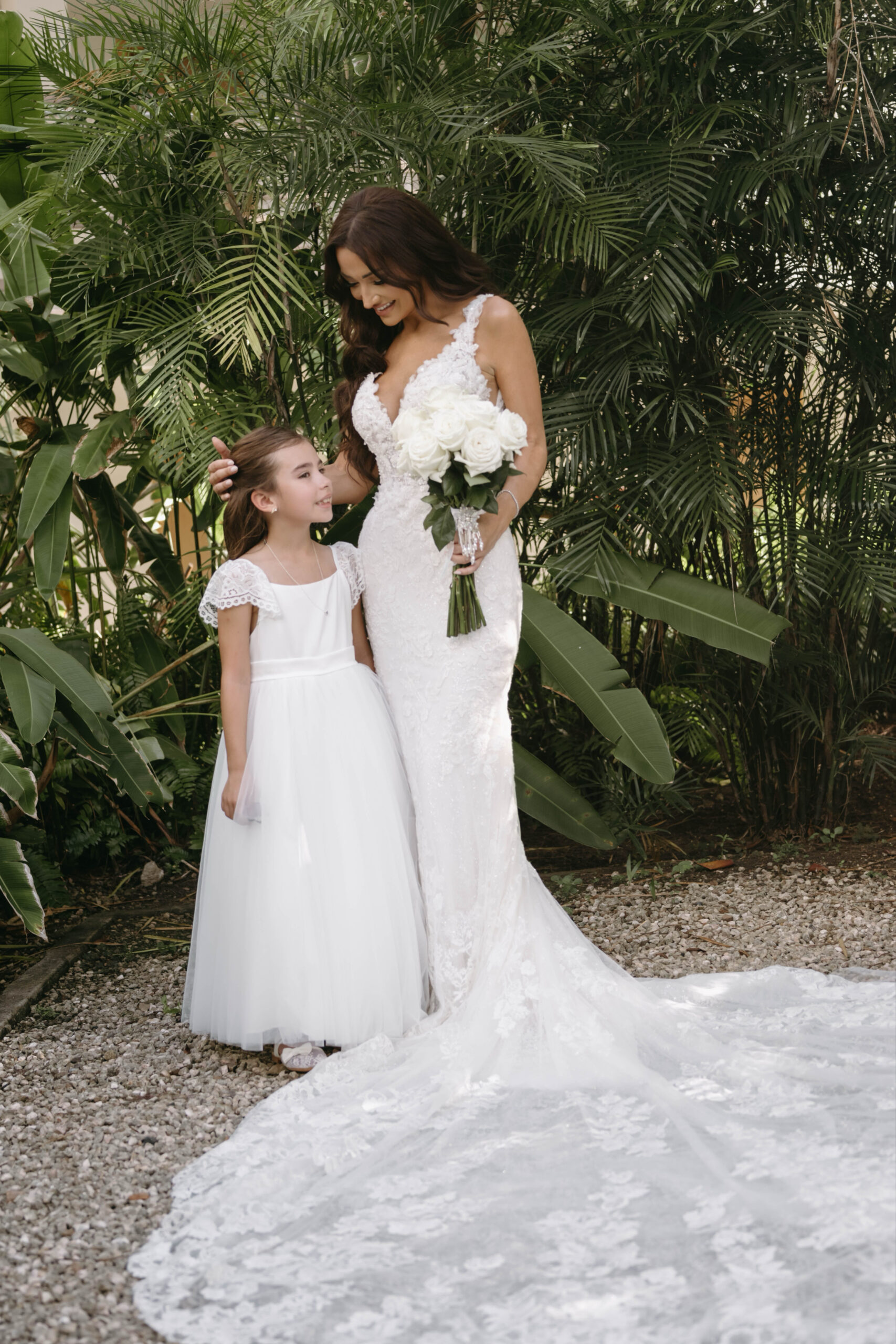 Bride and flower girl, both wearing white dresses and silver jewelry in front of green tropical backdrop.