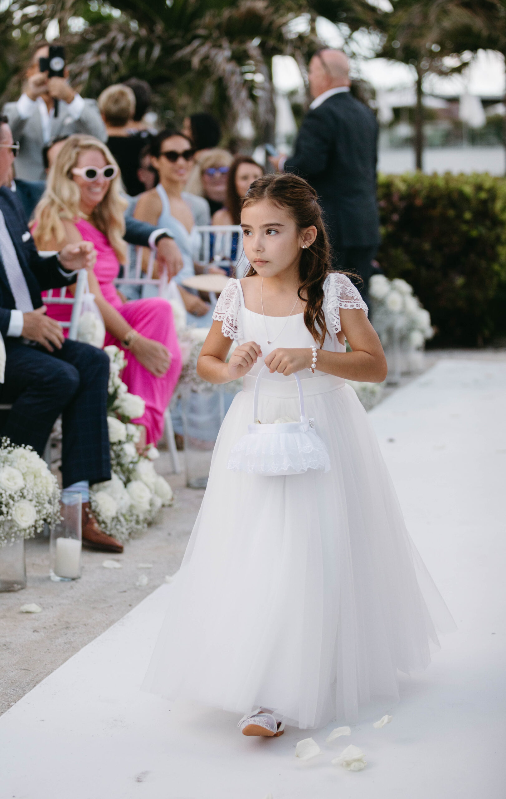 Flower girl walks down the aisle wearing Dune Jewelry and a white dress. She holds a white basket.