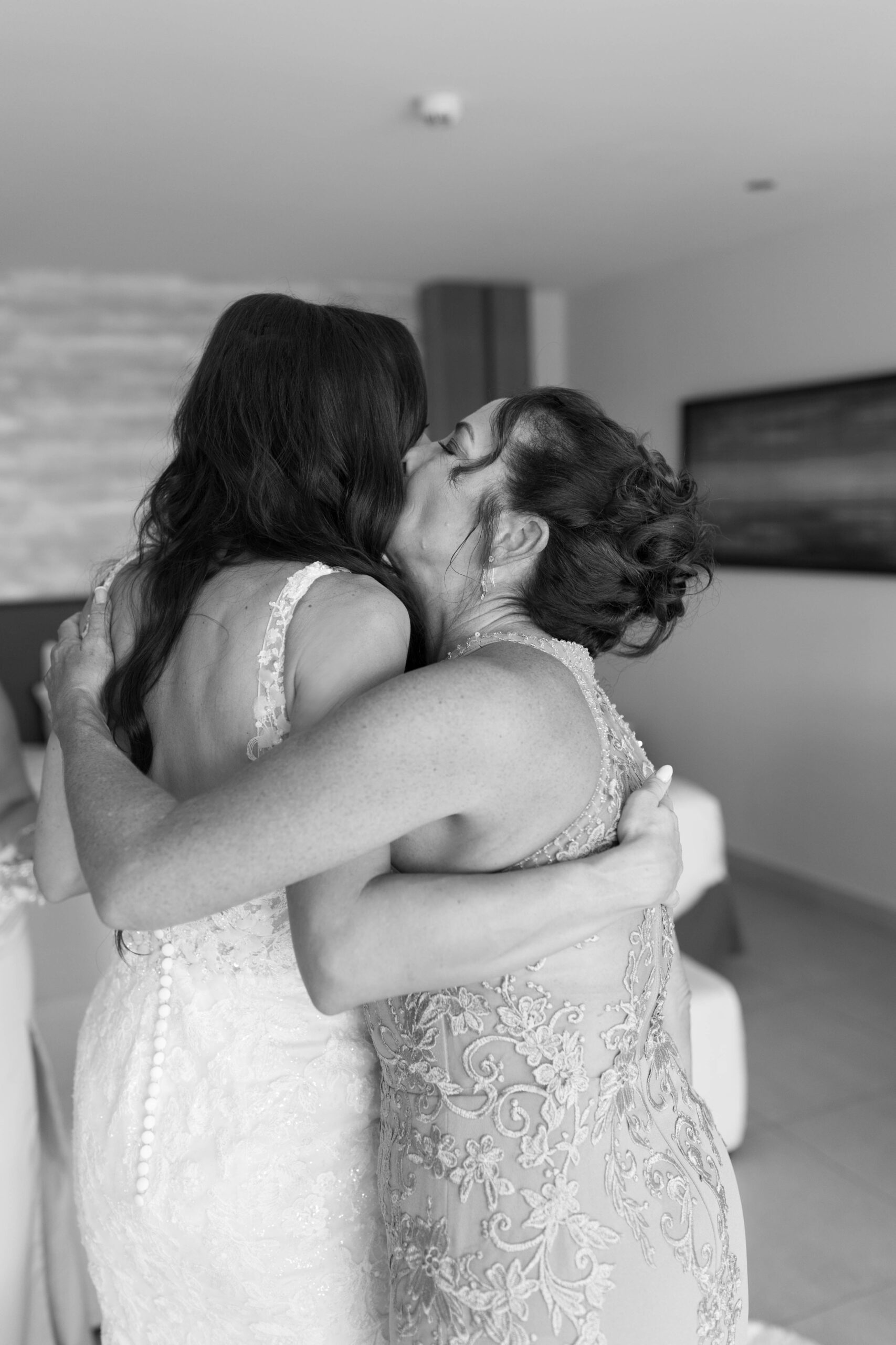 Black and white photo. Bride embraces her mother who gives her a kiss on the cheek. Their backs face the camera.