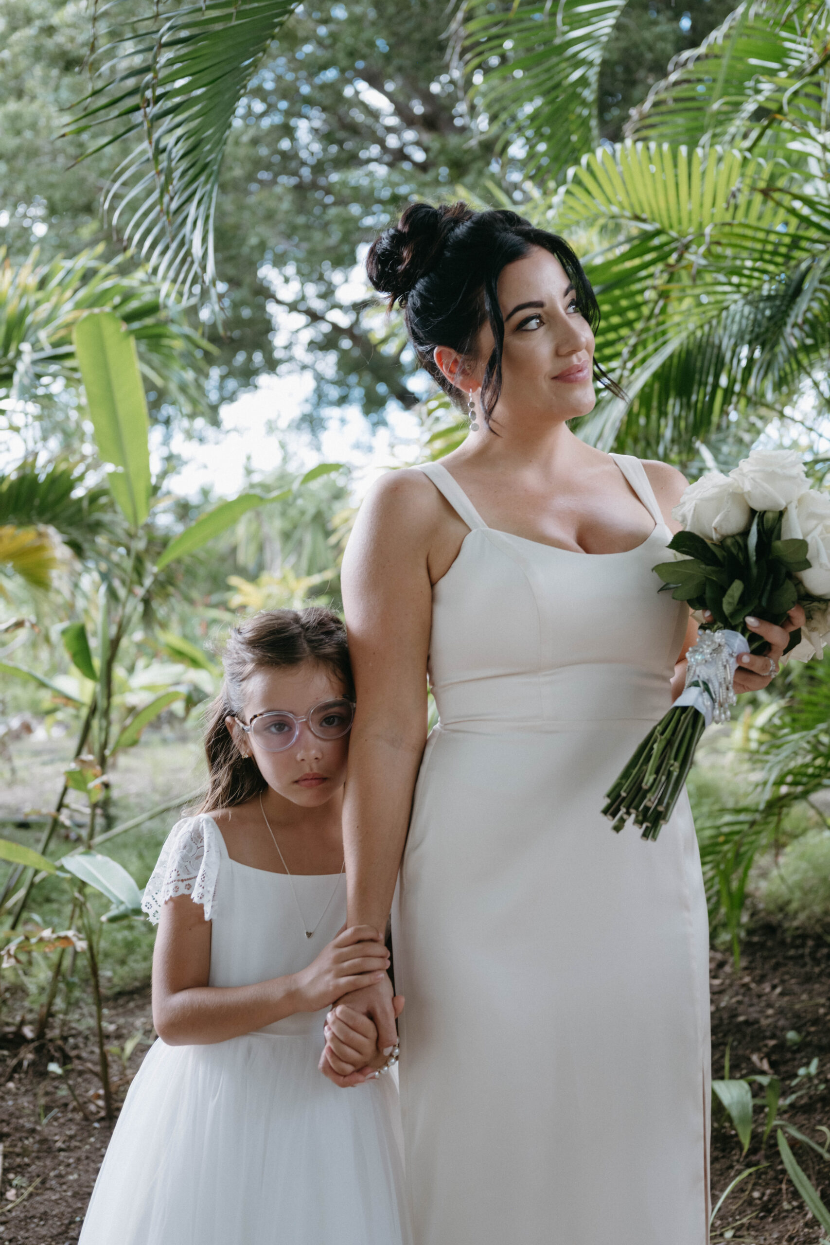 Flower girl and bridesmaid stand in front of green tropical background while bridesmaid holds white bouquet. Both wear white.