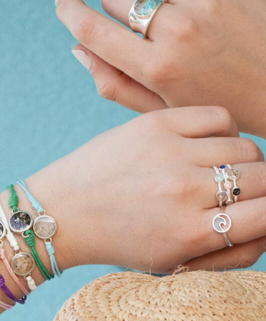 Two hands showcasing the Touch The World bracelets which include one sand and earth element each and colorful