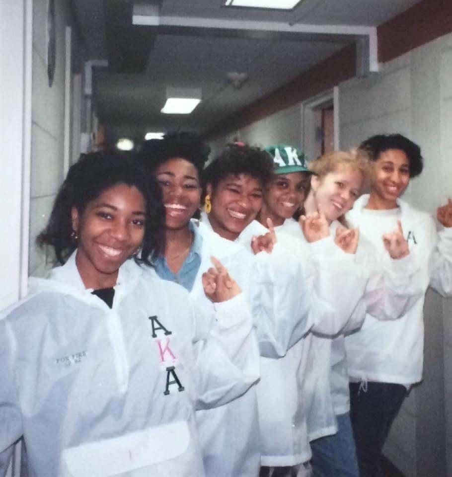 Six sorority girls holding up their pinkies and wearing white coats with letters AKA from their sorority on them in black and pink.