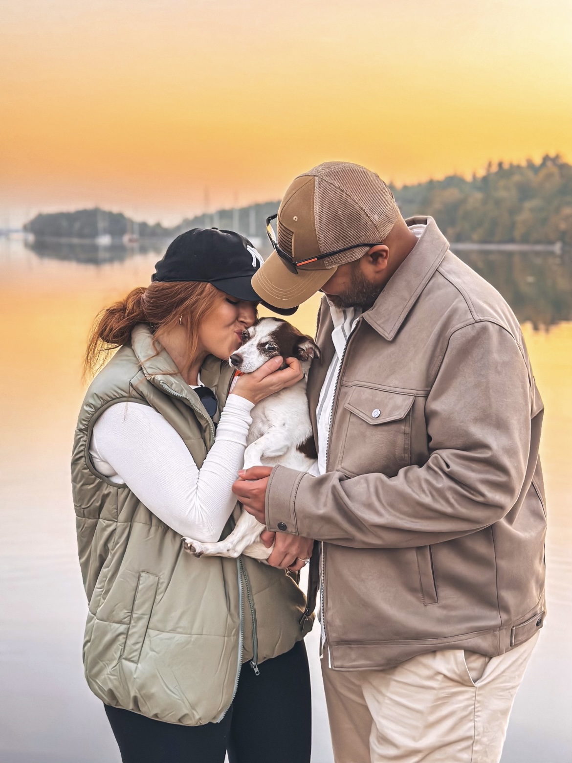 Jami and her partner holding and kissing their dog Mack with a scenic backdrop of a lack during sunset.